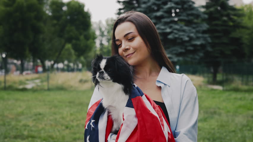 Happy Caucasian Woman Holding Her Japanese Chin Dog Wrapped in the USA Flag, Standing Outside in the Park. People and Dog Patriotism Concept | Shutterstock HD Video #1101745515