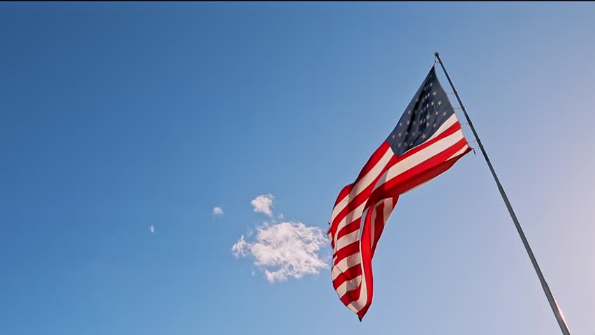 American USA flag on a flagpole waving in the wind against a clear blue sky on a sunny day. Close-up, bottom view, 4K. American Flag Waving In The United States Of America Shows Independence. Royalty-Free Stock Footage #1101747189
