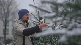 Man video blogger shoots video on his phone for social networks and subscribers on video services about the choice of a Christmas tree at the street before Christmas market in the snowy weather. 