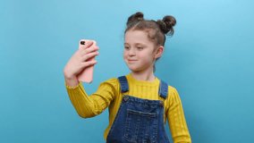 Happy little girl blogger taking selfie on smartphone holding in hand, communicating video call talking online with subscribers. Pretty child posing isolated on blue color background wall in studio