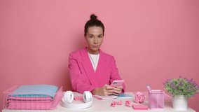 Young pleasant woman in pink blazer sitting at desk in pink studio office, makes a gesture with her hand pointing at the empty wall on right, business time concept, copy space, high quality video