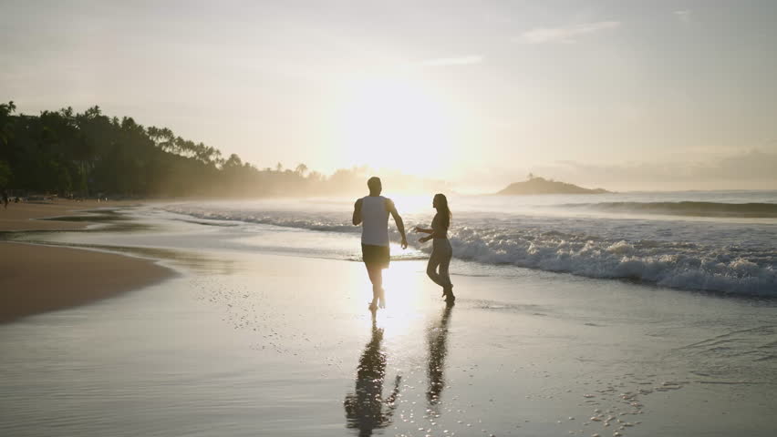 Silhouettes of a young happy couple holding hands and running on the beach together enjoying summer back view. Boyfriend and girlfriend having fun at the seaside hugging and kissing at sunrise.
