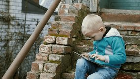 A handsome boy of 5 years old is watching a video or playing a mobile game on a tablet while sitting on the steps of a dilapidated, shabby, dilapidated building.