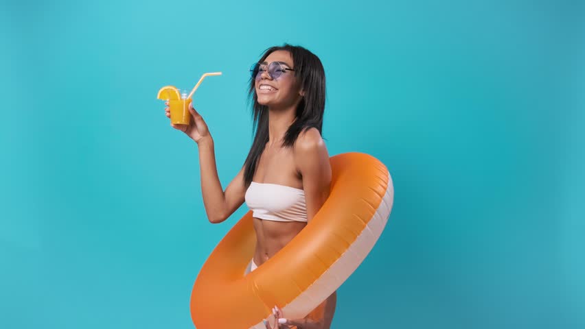 Young African American girl wearing swimsuit overall standing isolated on blue wall holding swim ring drinking fresh fruit smoothie smiling joyful