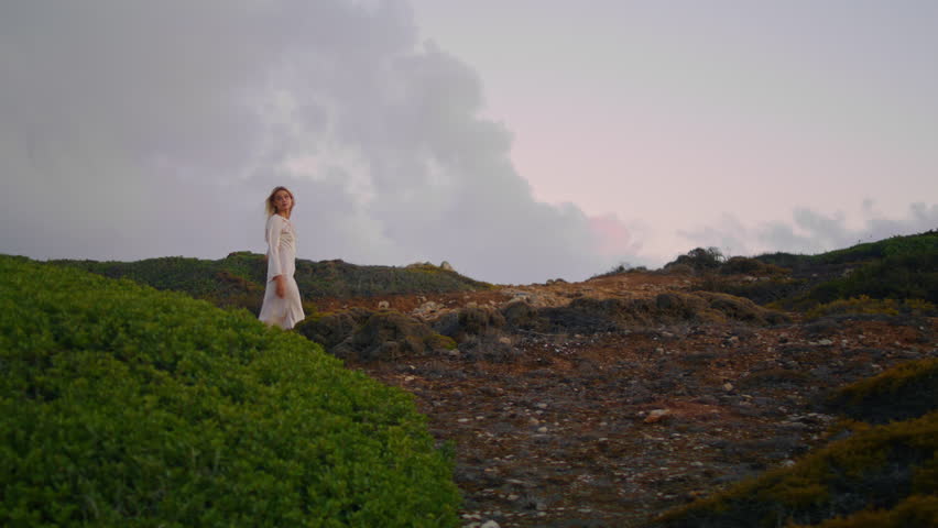 Serene model going nature cloudy sky background. Seductive girl wearing dress walking sunset grassy hill. Young happy lady looking distance relaxing green valley. Blonde woman spending evening alone