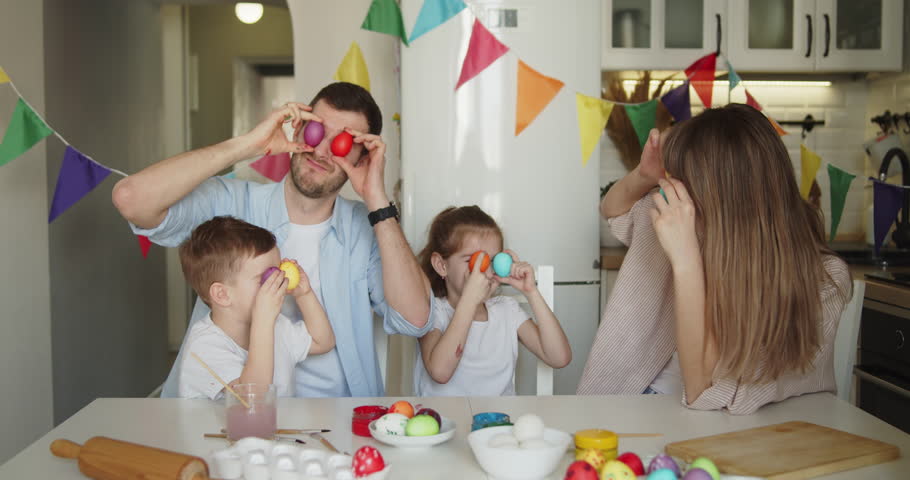 Joyful family of four painting Easter eggs together at the table in bright modern kitchen. Parents and children have fun playing with colored eggs. Family gathering in preparation for Easter.