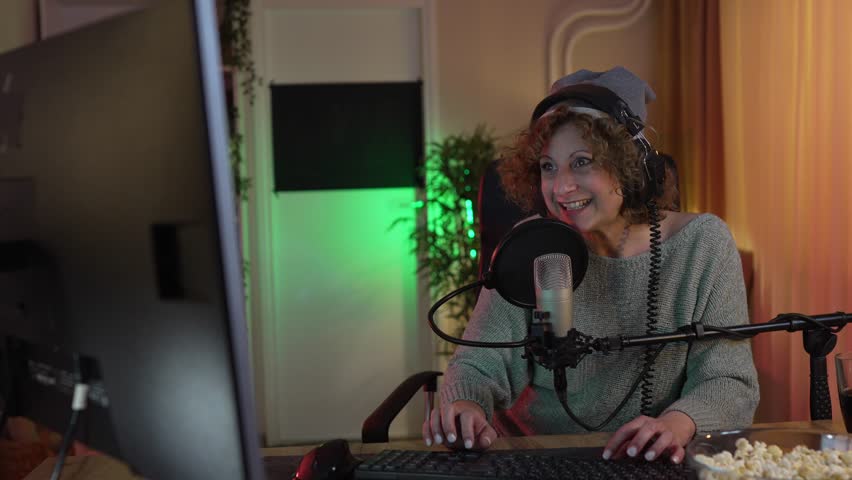 Mature woman play video games on pc computer while streaming to social media or internet online playtrough or walktrough video female gamer having fun at home wear headphone happy winning