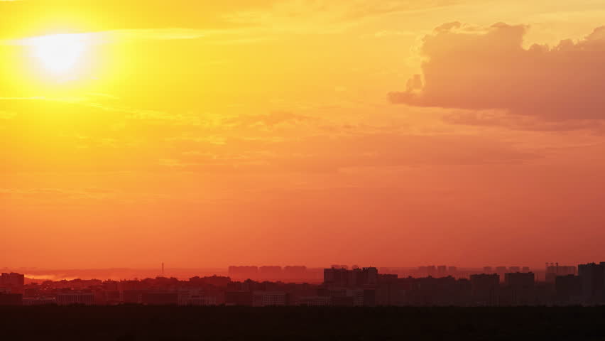 Big red sun over the roofs of city buildings, evening sunset. Timelapse of a cloudy sky and the sun setting behind the houses