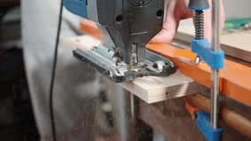 Carpenter caucasian man with an electric jig saw in his hands cuts piece from sheet of plywood fixed on table and sawdust from fret saw flies to the sides. Master saws for furniture. Close up video.