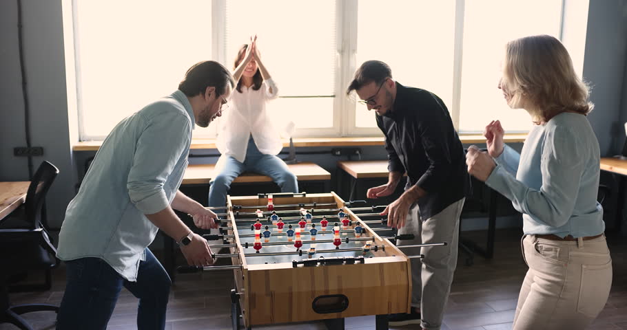 Excited office employees having fun, playing table soccer, laughing, smiling, winning with high five. Different aged workers giving support to active tabletop football players, shouting Royalty-Free Stock Footage #1101777783