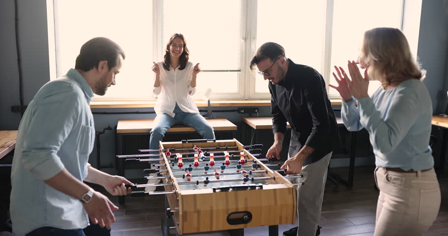 Excited office employees having fun, playing table soccer, laughing, smiling, winning with high five. Different aged workers giving support to active tabletop football players, shouting