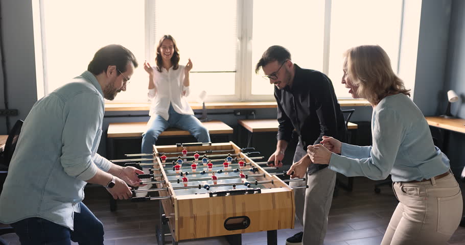 Excited office employees having fun, playing table soccer, laughing, smiling, winning with high five. Different aged workers giving support to active tabletop football players, shouting
