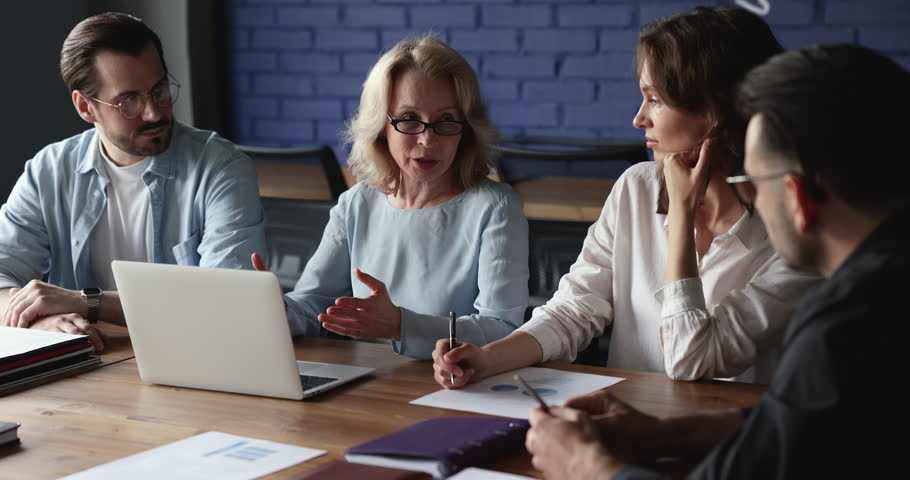 Senior mentor woman sitting at table with laptop, talking to team of listening interns, employees, discussing work project, learning tasks, asking questions, sharing ideas, brainstorming Royalty-Free Stock Footage #1101777807