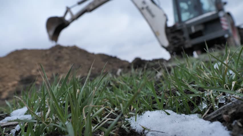 View from the ground. Snow on green grass. On an blurry background, an excavatar digs a trench. Close-up
