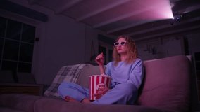 Young caucasian woman watching sad 3d movie on a projector at home. Emotional woman in blue suit sitting on couch with popcorn. High quality 4k footage