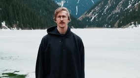man in glasses stands with background of a lake and mountains, puts on a hood. High quality FullHD footage