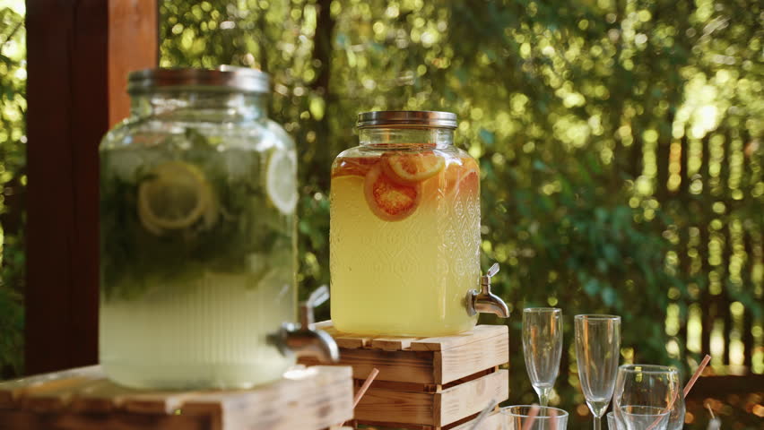 Two glass lemonades with cooling fruit drinks stand on wooden stand made of planks. Glass jars with tap, with cocktail stand in green yard of house. There are empty glasses near drink bar. | Shutterstock HD Video #1101821927
