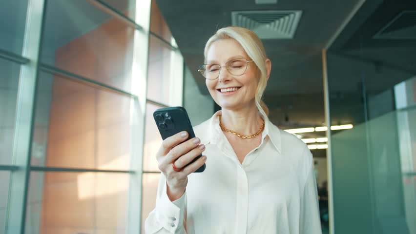Smiling mature businesswoman holding smartphone walking in office. 50s aged manager CEO using cell phone mobile apps. Digital technology applications and solutions for business corporate, slow motion | Shutterstock HD Video #1101825695