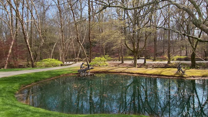 Footage of a green pond with statues of children on the banks with lush green grass, moss, red trees and lush green plants at Gibbs Gardens in Ball Ground Georgia USA