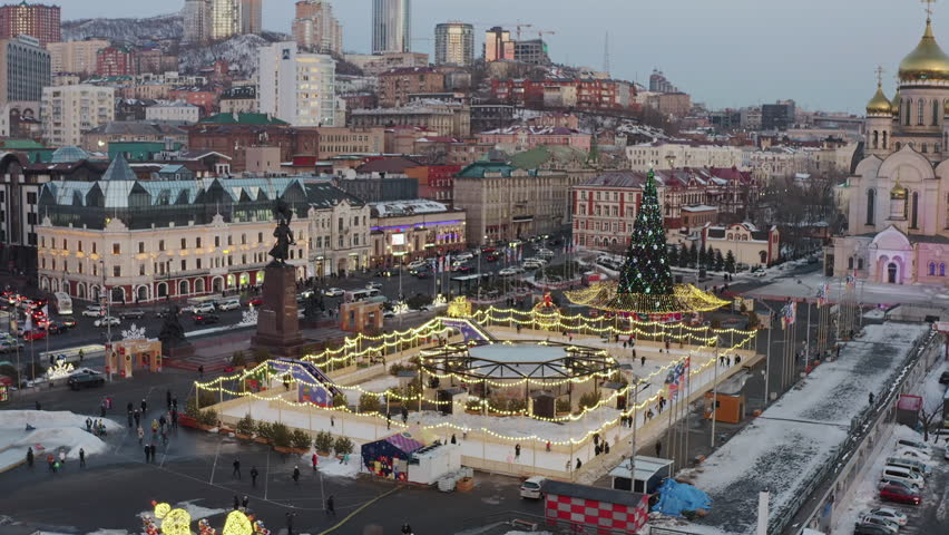 Aerial view of the central square of the city of Vladivostok with a skating rink and ice slides at sunset in winter. People rest and rush home after work. Traffic jams