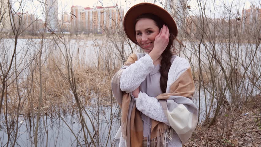 A young woman in a light jacket with a scarf puts on a hat near the water against the background of dry branches. A woman in her thirties is cheerful. Woman with beautiful long hair