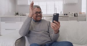 Video call, waving and greeting on a phone with a senior man sitting in his living room at home. Happy, smiling and positive mature male gesturing and giving thumbs up while talking and laughing