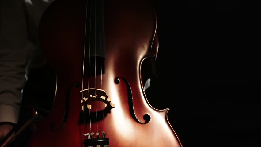 Professional musician preparing to start playing cello putting his left hand on the fingerboard and touching strings with a bow in his right hand close up in a dark room | Shutterstock HD Video #1101849575