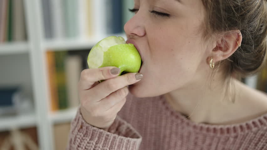 Young beautiful hispanic woman eating green apple at library university | Shutterstock HD Video #1101850825