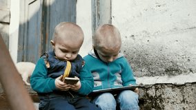 Portrait of children looking at mobile gadgets. The brothers watch videos or play video games on their mobile phones and tablets while sitting on the steps of a dilapidated building.