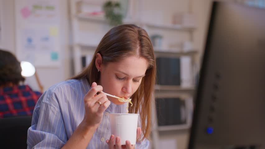 Caucasian businesswoman eatting noodles while work in office at night. Attractive professional female employee sitting on table, feeling hungry and starving then having dinner in corporate workplace.