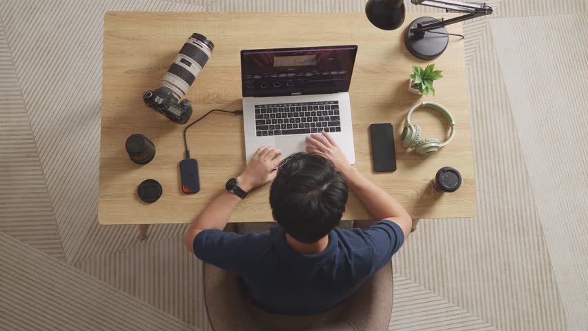 Top View Of A Male Color Grading Sitting In The Workspace Using A Laptop Next To The Camera Editing The Video At Home
