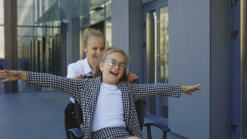 Two Excited Little Businesswoman Having Fun Riding on Office Chairs Outside Near Office Building. Business Kids Take a Break, Enjoy Playing Games, and Laughing Together in Modern Business Center | Shutterstock HD Video #1101853029