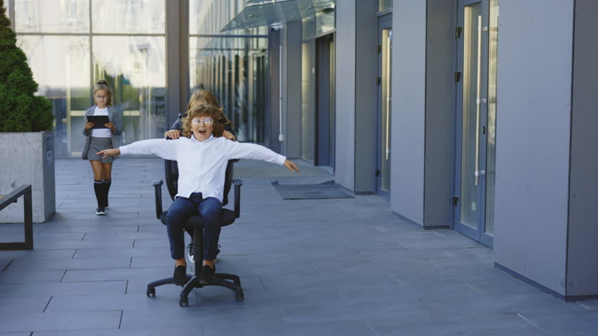 Excited Little Businessperson Having Fun Riding on Office Chairs Outside Near Office Building. Business Kids Take a Break, Enjoy Playing Games, and Laughing Together in Modern Business Center | Shutterstock HD Video #1101853035
