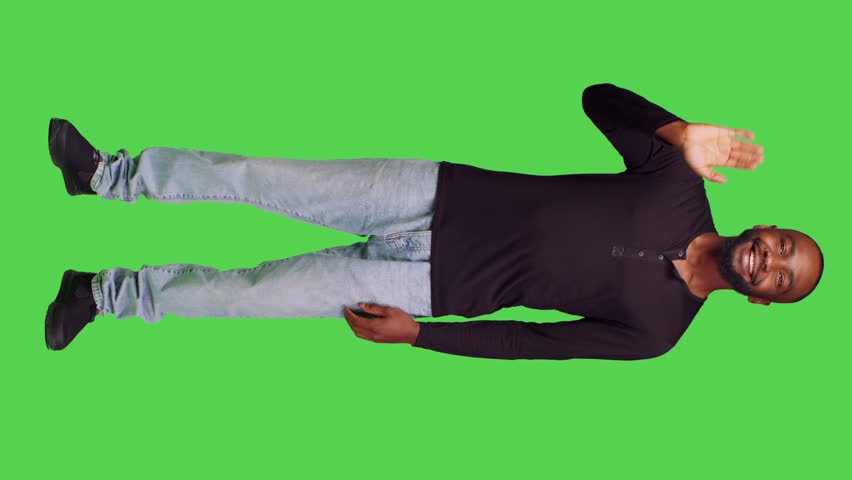 Vertical video: Front view of african american guy waving at people in studio, saying hello or goodbye on camera. Young model smiling and greeting someone standing over full body green screen