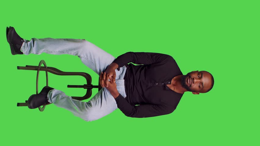 Vertical video: Front view of african american man on chair posing with confidence, sitting over full body green screen backdrop. Young male model feeling optimistic and waiting for something in Royalty-Free Stock Footage #1101853521