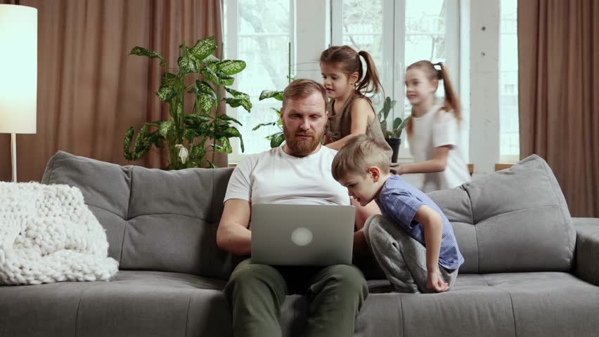 Too noisy. Man, father sitting on couch at home and working remotely with his little children loudly playing around him. Concept of fatherhood, childhood, freelance job, remote work, emotions Royalty-Free Stock Footage #1101857775