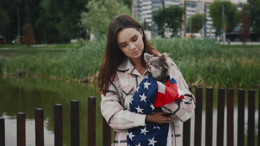 Portrait of the caucasian woman owner smiling holding her small chihuahua wrapped in the usa flag standing outside in the park looking at the camera. People and dog patriotism concept | Shutterstock HD Video #1101865307