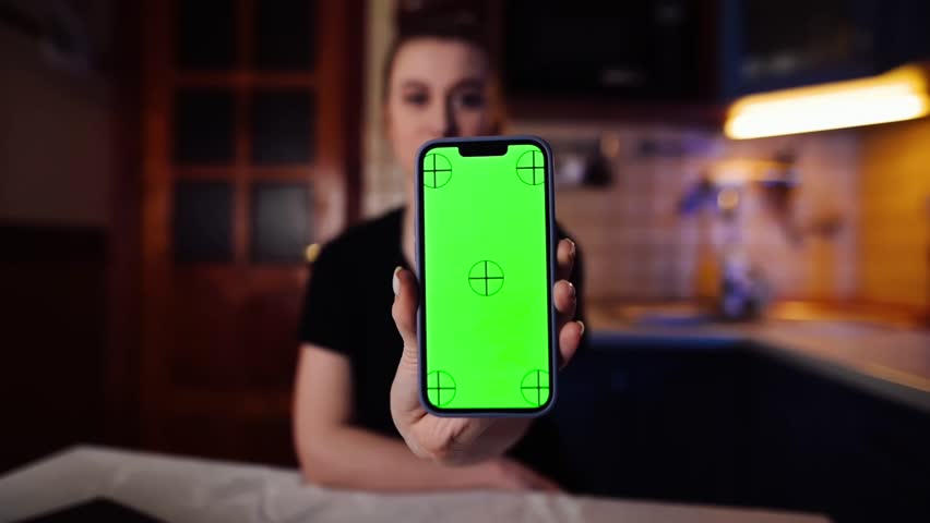 A smiling young girl sits in the kitchen and shows a smartphone with a green screen layout. Advertising of modern mobile applications. Close up | Shutterstock HD Video #1101867307