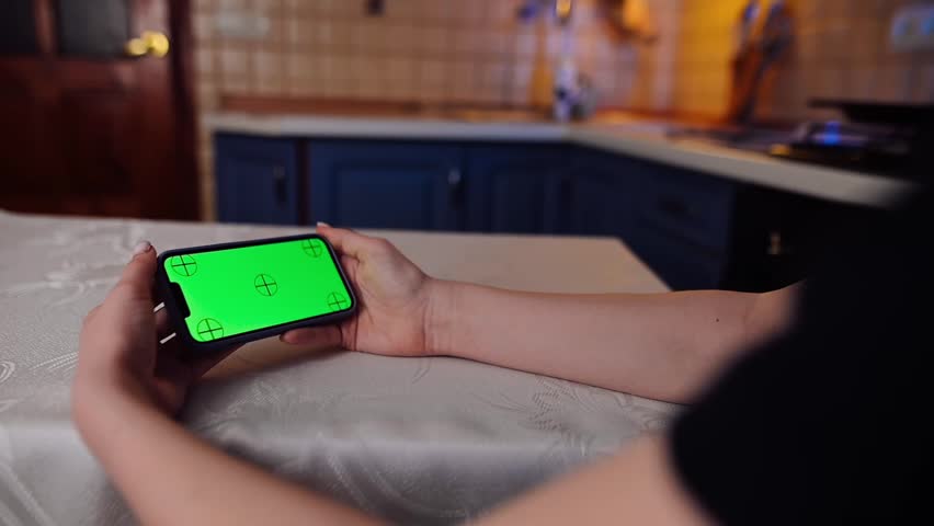 A woman looks at a horizontal phone with a green screen, sitting at the table in the kitchen in the evening. A girl is surfing the internet. Technological content. | Shutterstock HD Video #1101867311