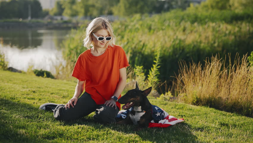 Portrait of the Beautiful Stylish Woman Sitting on the Grass with the Mini Bull Terrier Is Wrapped in the USA Flag in the Park. Patriotic Dog Lying on the Grass Near Girl Looking at Camera | Shutterstock HD Video #1101867483