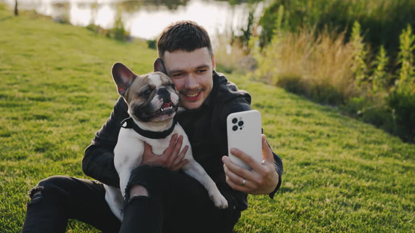 Handsome Smiling Caucasian Man Is Sitting on the Grass with His Brooklyn Bulldog and Doing Selfie on Smartphone. Guy Making Photo Memories with His Pet. Technology and People Dogs Friendship | Shutterstock HD Video #1101869189
