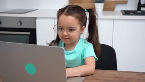 Little girl with glasses using a laptop, doing homework in the kitchen at home.
