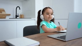 Cute little girl writes in a notebook during a preschool online lesson at home.