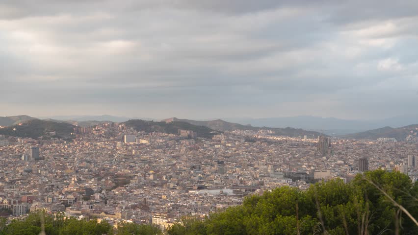 Timelapse over Barcelona city; epic view on Sagrada Familia from Montjuïc Castle; clouds and sunlight over city in Autumn during sunset; hills, houses and massive sprawling big city covering horizon