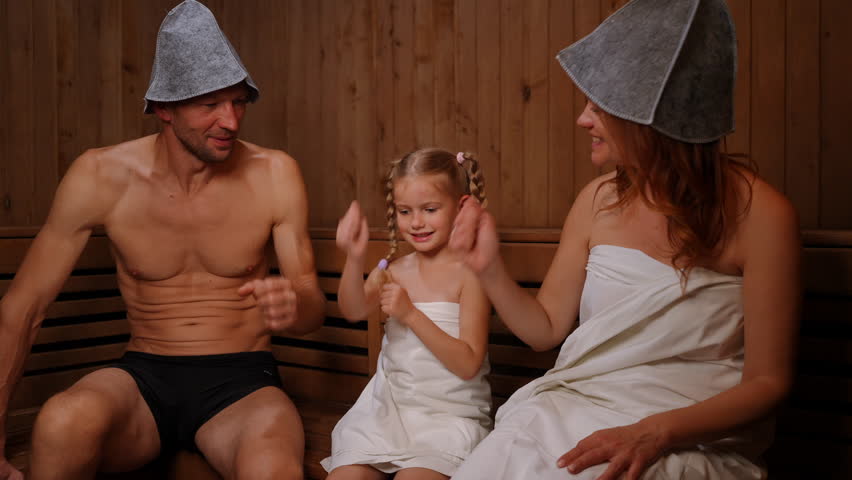 Joyful daughter playing rock-paper-scissors with parents sitting on wooden bench in Finn sauna. Portrait of cheerful laughing Caucasian girl having fun with father and mother in SPA indoors