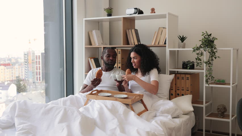 African american family man and woman eating crescent rolls and drinking black coffee while lying on bed in sunny morning. Joyful married couple sending message of true love through daily care.