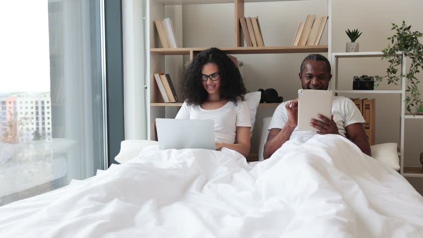 African american romantic partners using electronic devices while lounging on wide bed near panoramic window. Beautiful woman with laptop and relaxed man with tablet enjoying leisure time at home.