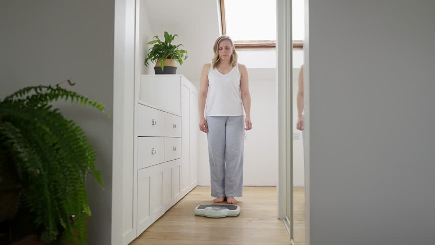 Menopausal Mature Woman Concerned With Weight Gain Standing On Scales In Bedroom At Home Royalty-Free Stock Footage #1101896367