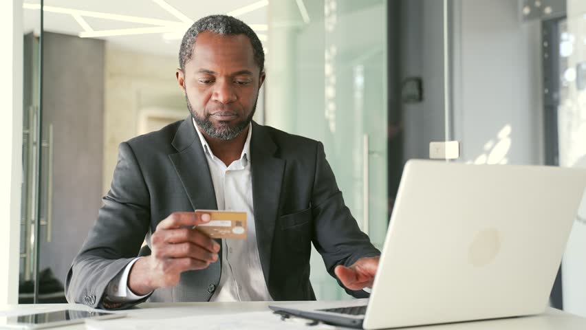 Upset african american man holding credit card and looking at laptop screen while sitting in office. Businessman is angry and frustrated because he faced a fraud. He had money stolen from his account Royalty-Free Stock Footage #1101897485