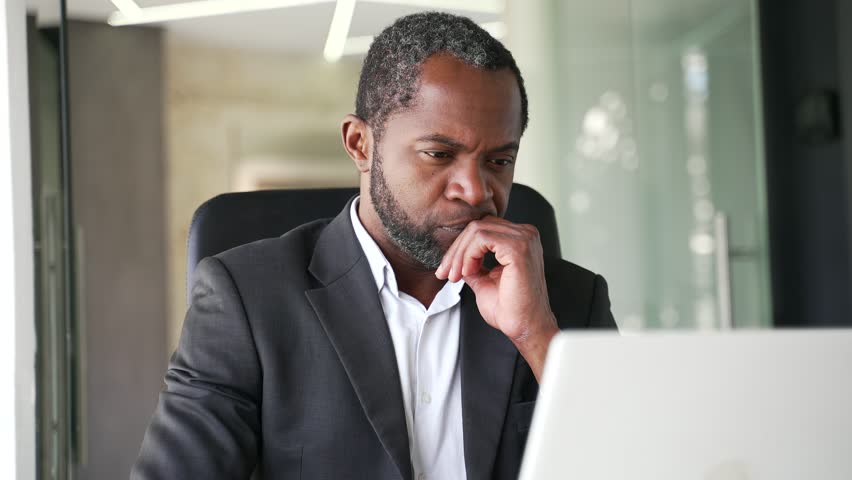 Close up of a thoughtful mature african american man in a formal suit sitting at a workplace in a office using a laptop. Concentrated middle aged businessman is thinking about a serious business deal Royalty-Free Stock Footage #1101897493